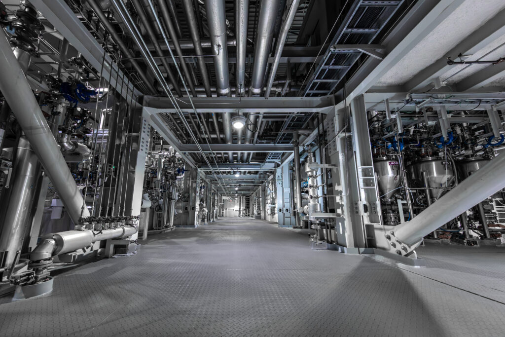 Interior corridor of a battery electrolyte manufacturing facility with stainless steel piping, reactors, and industrial process equipment