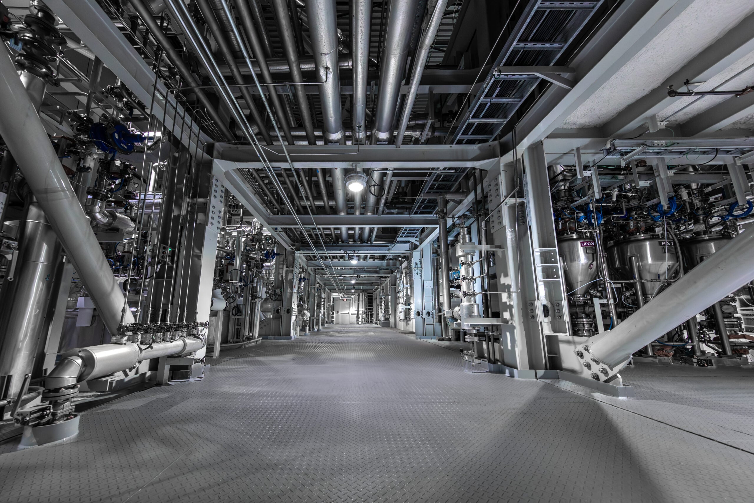 Interior corridor of a battery electrolyte manufacturing facility with stainless steel piping, reactors, and industrial process equipment