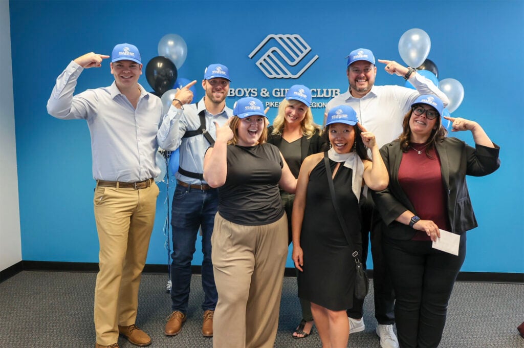 Seven staff members pose for a photo in front of a blue wall with a Boys & Girls Club sign, wearing matching blue caps and balloons in the background.