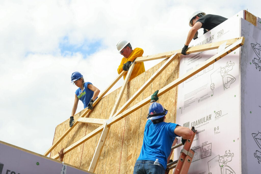 Construction workers in hard hats assemble a wooden frame on a building under construction at height.