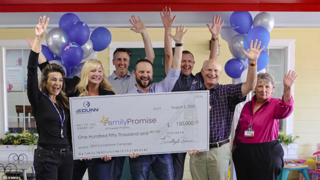 Group of eight adults celebrating and waving hands while holding a large ceremonial check, balloons in background.