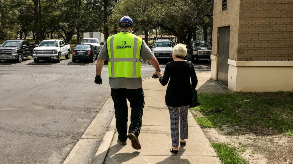 Construction worker in a neon safety vest and hard hat walking with an older woman along a sidewalk beside parked cars. They are holding hands as they walk toward a brick building in a parking lot.