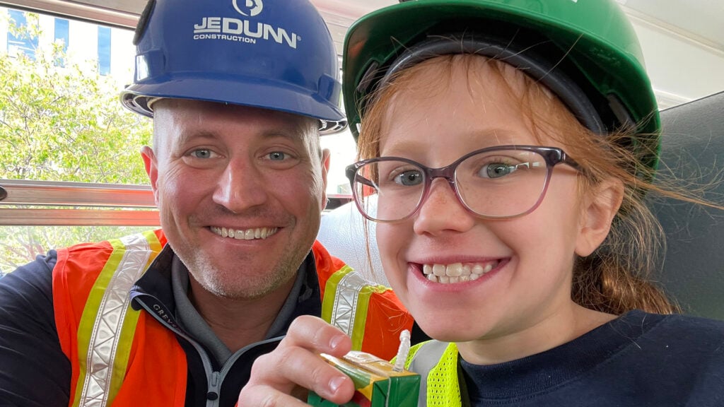Smiling man and girl in safety vests and hard hats taking a selfie; blue helmet reads 'Jedunn Construction'.