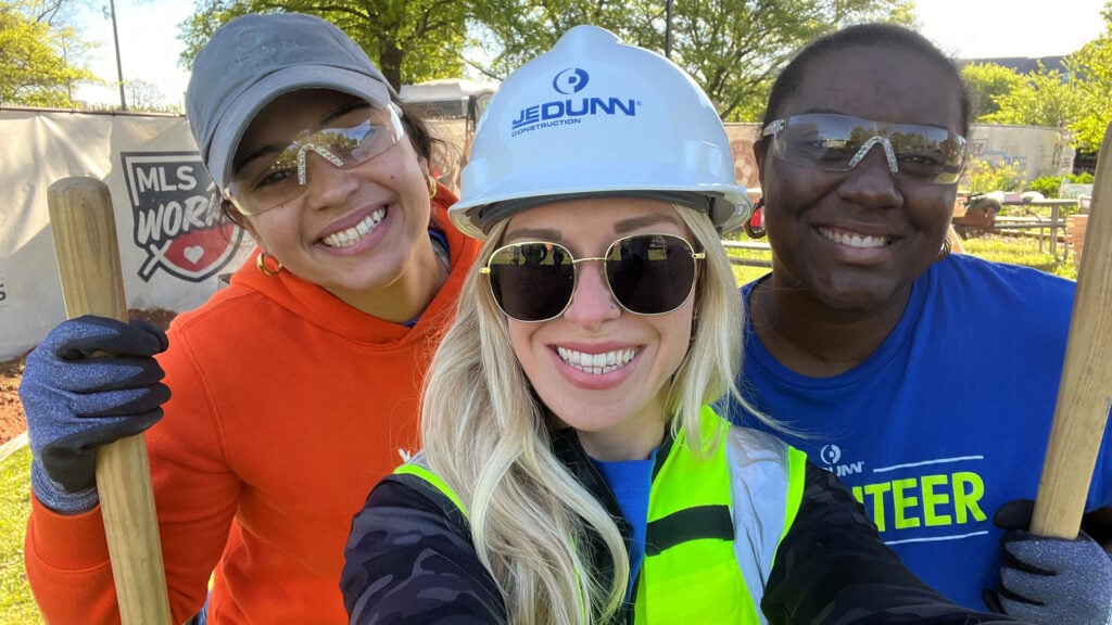 Three smiling volunteers posing for a selfie at a community build site, wearing safety gear (hard hat, reflective vest, gloves) and holding shovels.