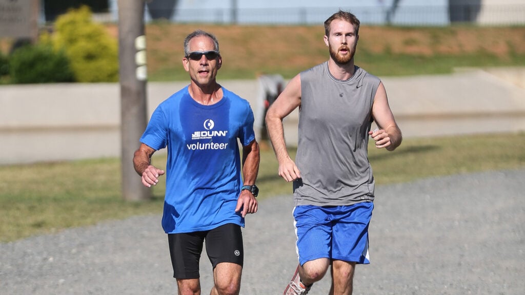 Two men are running side by side on a gravel path; one wears a blue 'Volunteer' shirt and the other a gray sleeveless shirt.