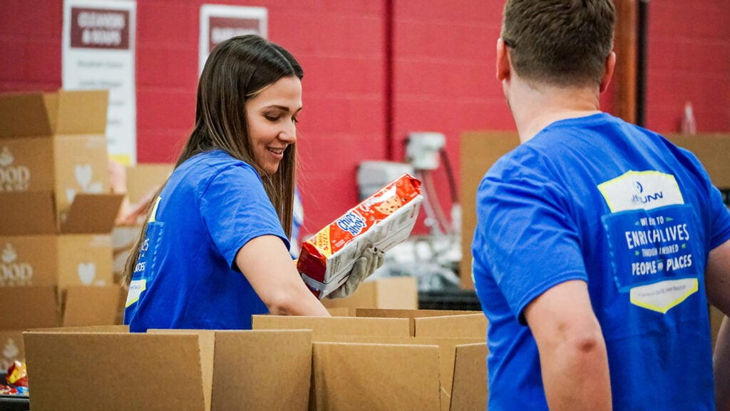 Two volunteers in blue shirts packing boxes in a warehouse, with one woman smiling while placing a snack pack into a box.
