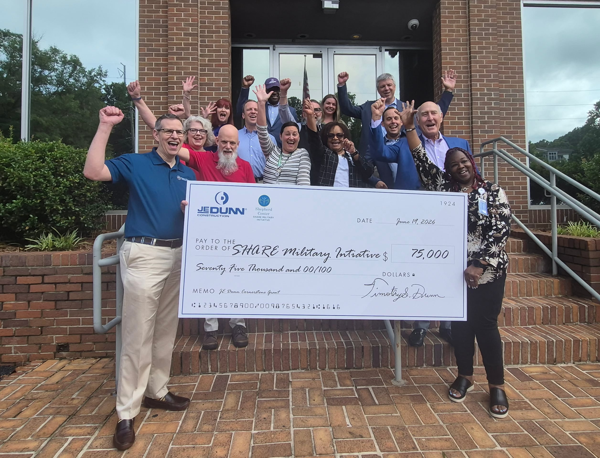 Group of diverse adults smiling and raising fists on brick steps, holding a large ceremonial check with logos and a date visible (June 19, 2026).