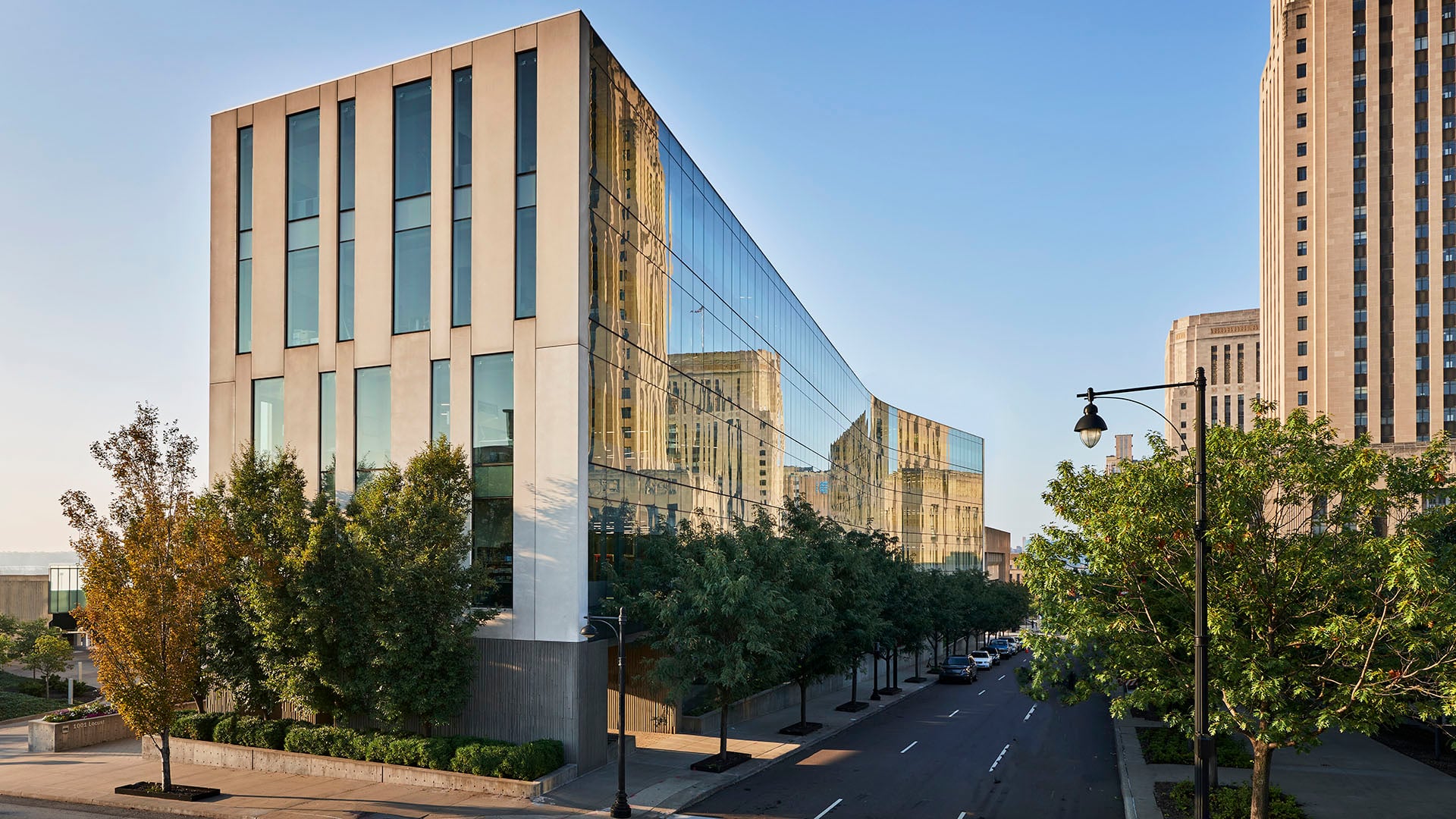 Modern glass-and-concrete office building with a curved reflective facade, reflecting surrounding towers on a sunny city street with trees.