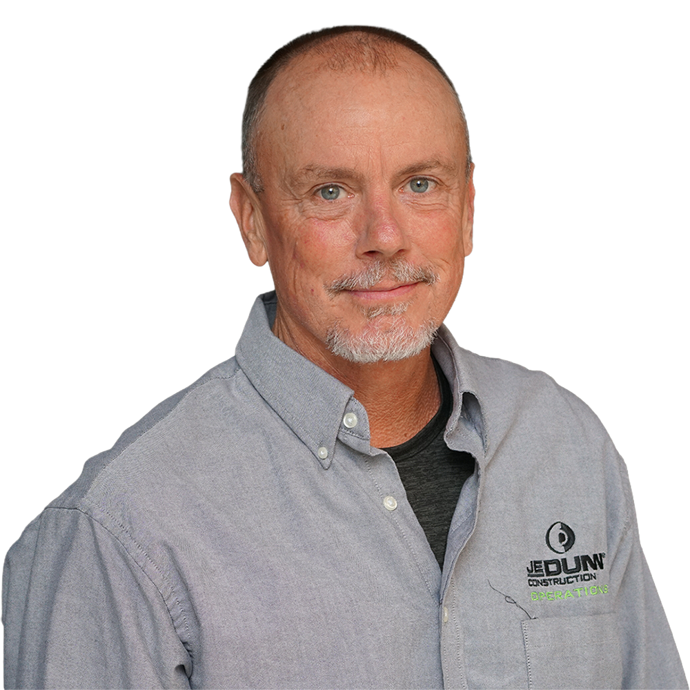 Smiling man in a gray button-down shirt with Jed Dunn Construction Operations logo, shown from the chest up against a dark background.