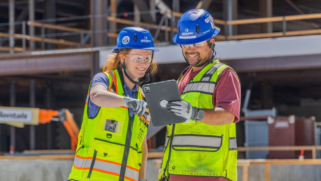 Two construction workers in hard hats and neon vests review information on a tablet at a building site.