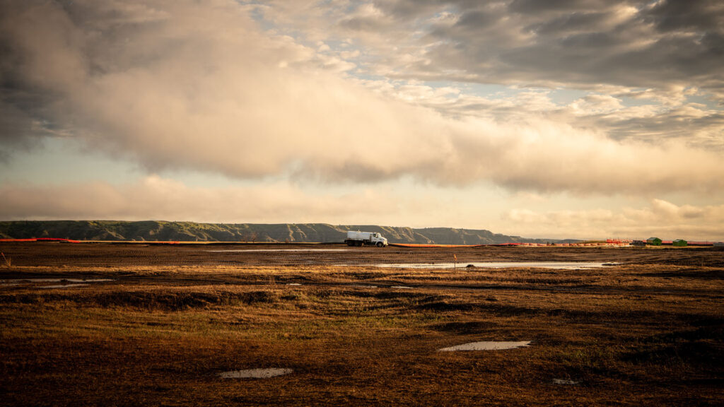 White tanker truck driving along a muddy field under a dramatic cloudy sky with low hills in the distance.
