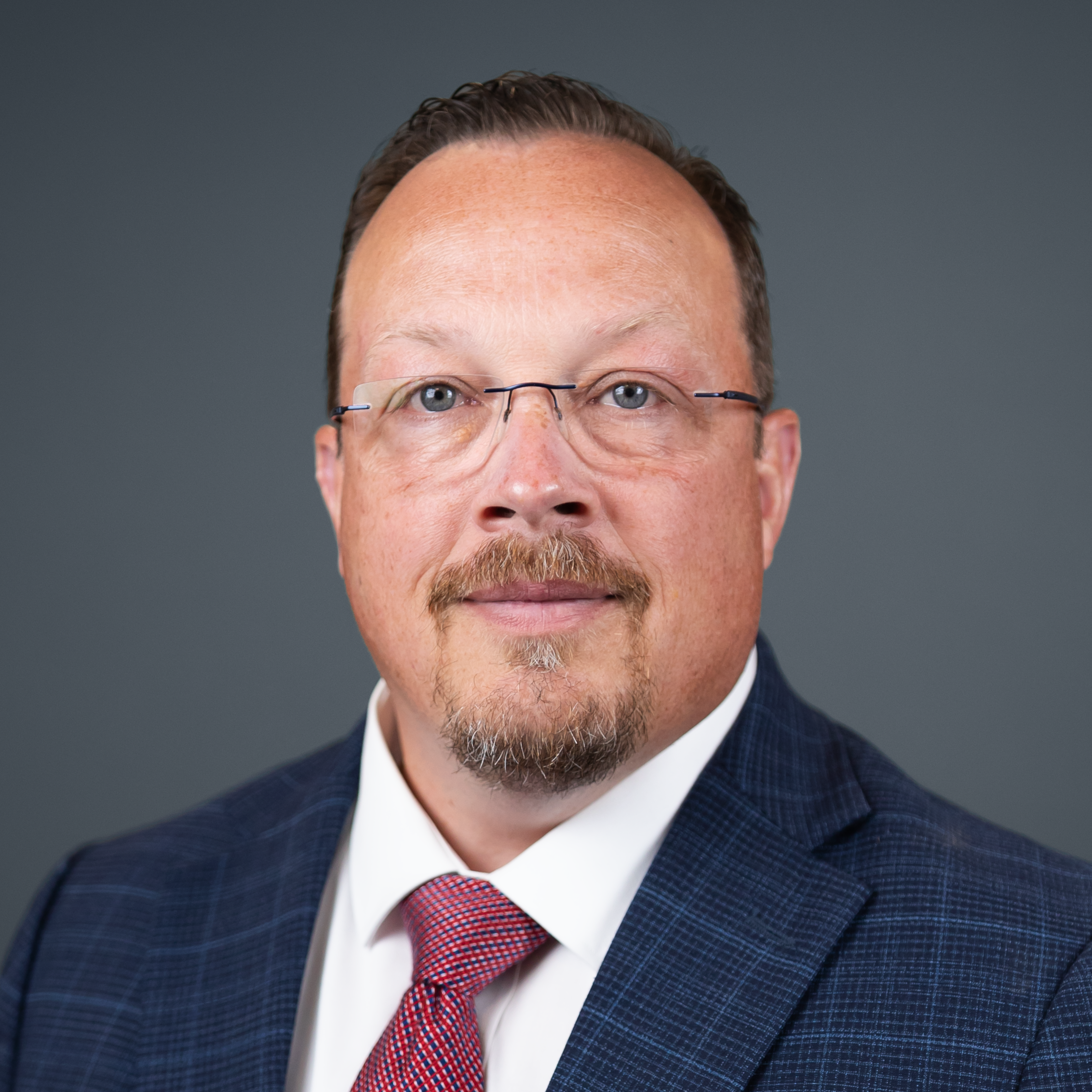 Close-up of a man in a navy check suit and red tie, wearing glasses, against a gray backdrop.