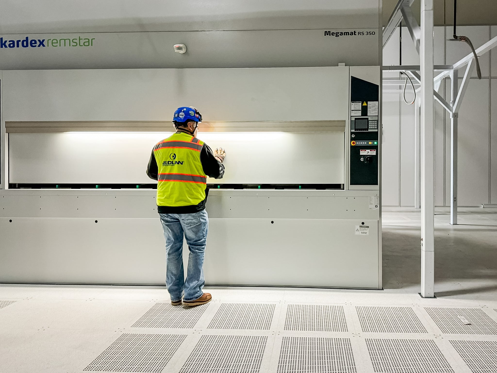 Worker in a high-visibility safety vest and blue hard hat stands at a large Kardex Remstar Megamat RS 350 automated storage system in a clean industrial setting.
