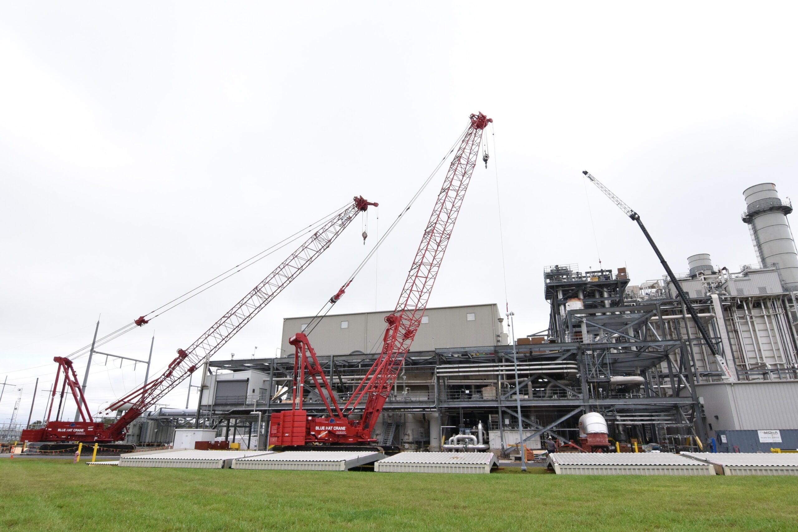 Construction site with large red cranes and an industrial building; green lawn in the foreground under a cloudy sky.