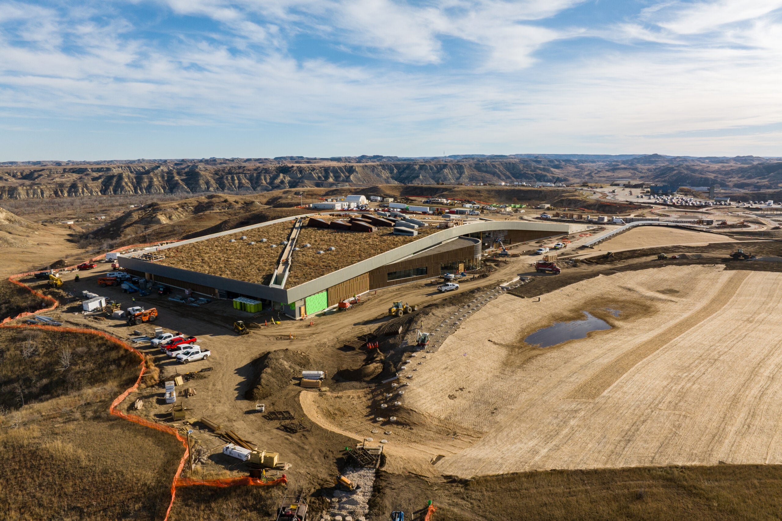 Aerial view of a large construction site with a curved, elongated building and construction equipment, set in a dry, hilly valley distance hills in the background