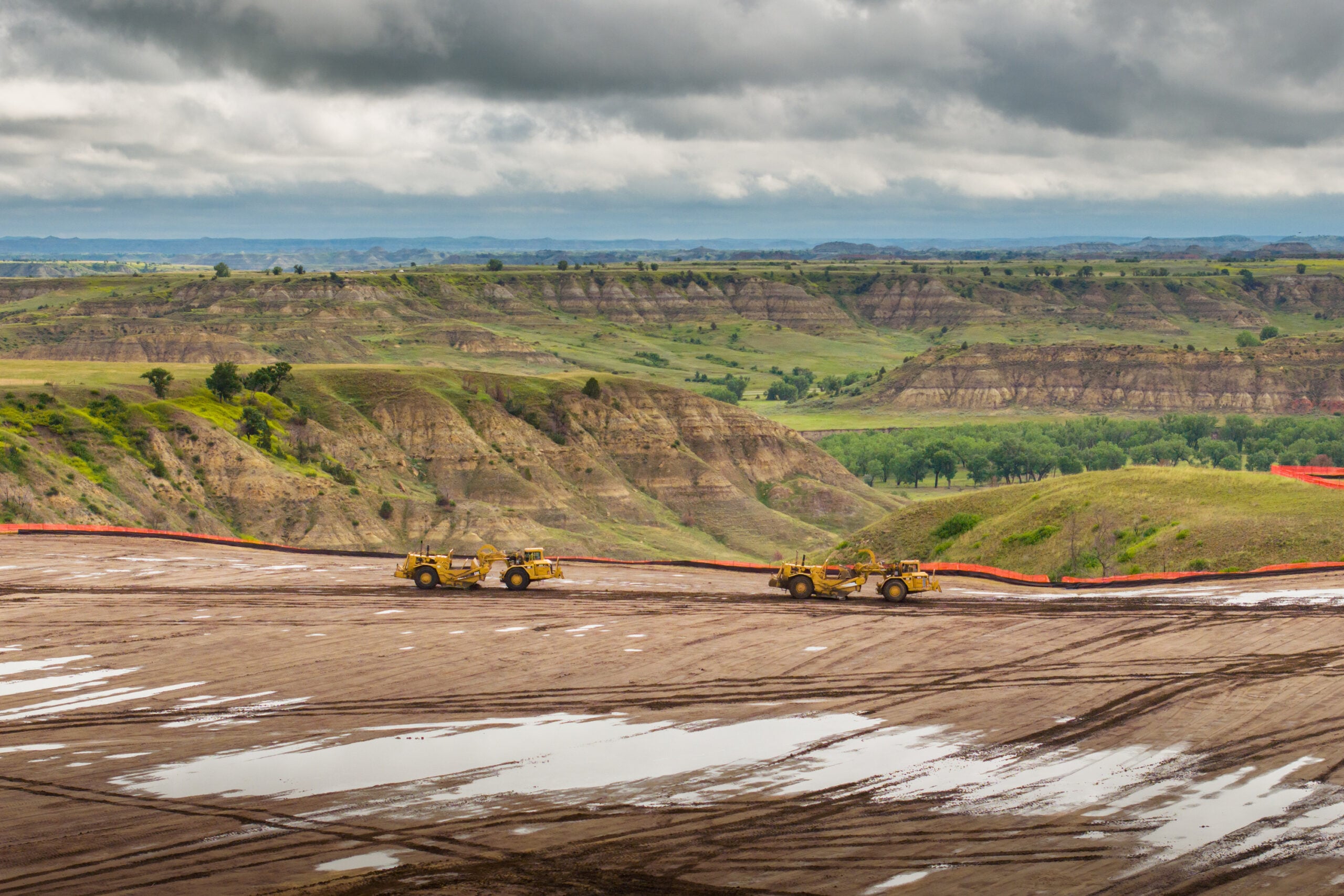 Construction site in a valley with dirt roads, yellow heavy equipment, and orange safety barriers under cloudy skies.