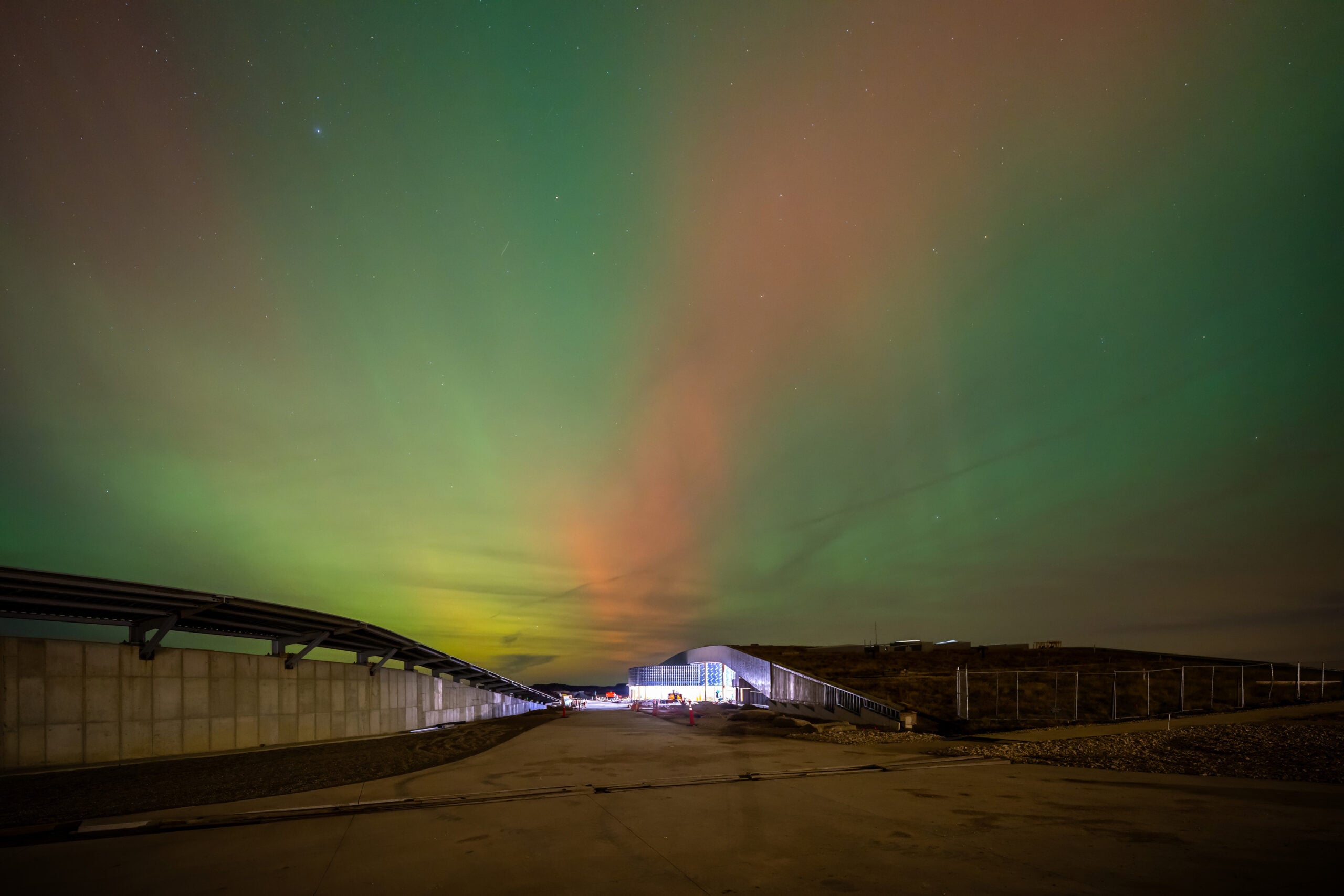Northern lights—green and red—illuminate a night-time industrial yard with a curved road and fences visible in the foreground.