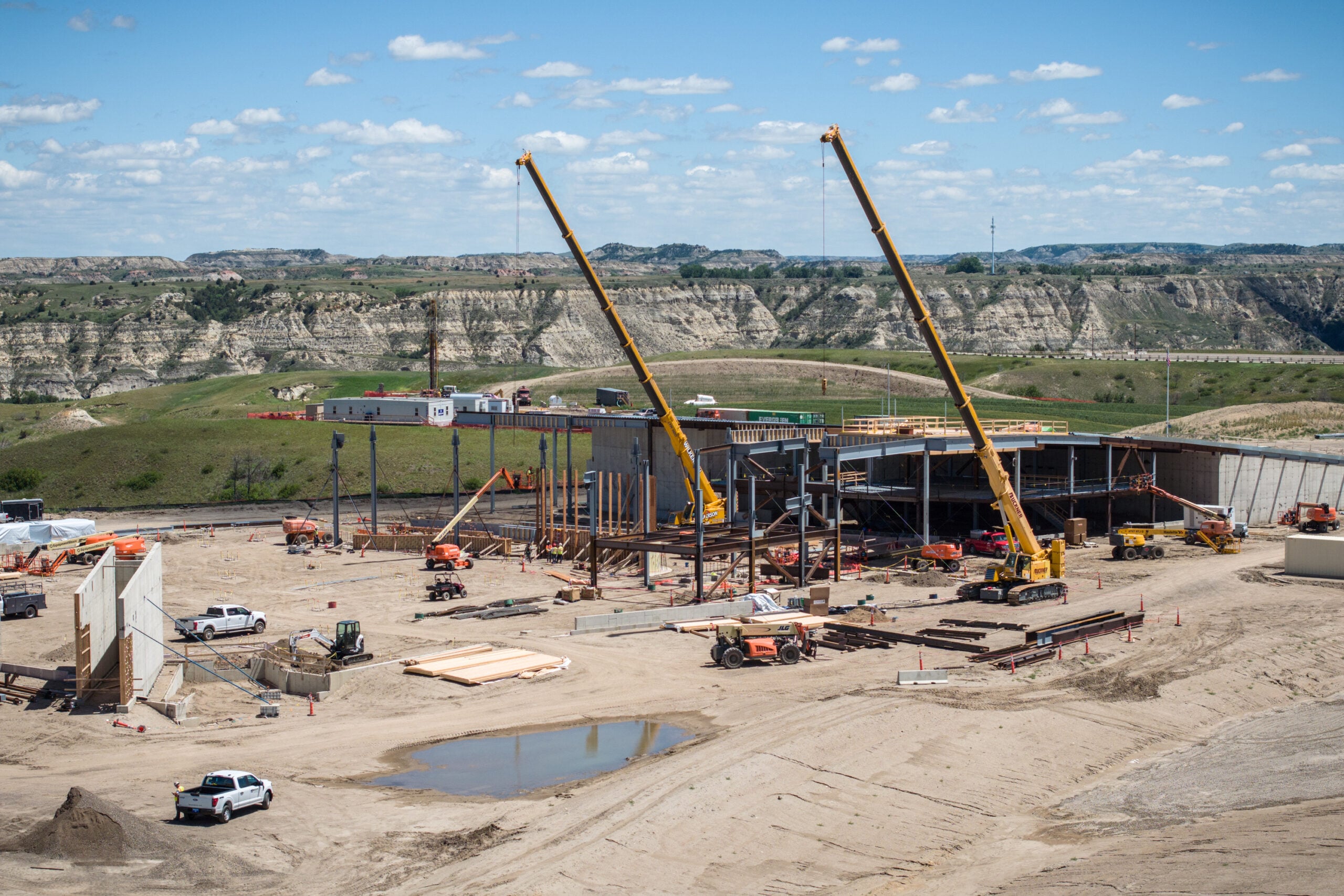 Construction site with two tall yellow cranes over a steel framework and scattered equipment, hills in the distance.