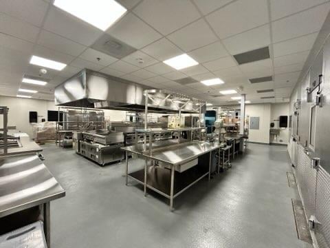 Large commercial kitchen with stainless steel prep tables, ovens, and an overhead exhaust hood; bright lighting and a worker visible in the distance.