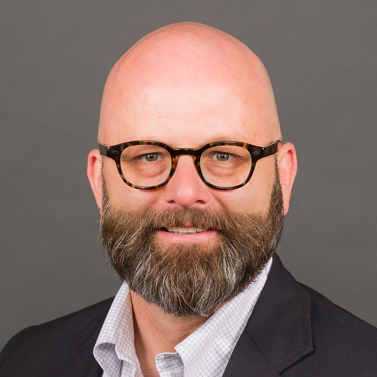 Portrait of a bald man with a full beard and tortoiseshell glasses, wearing a dark blazer and light checkered shirt against a gray background.