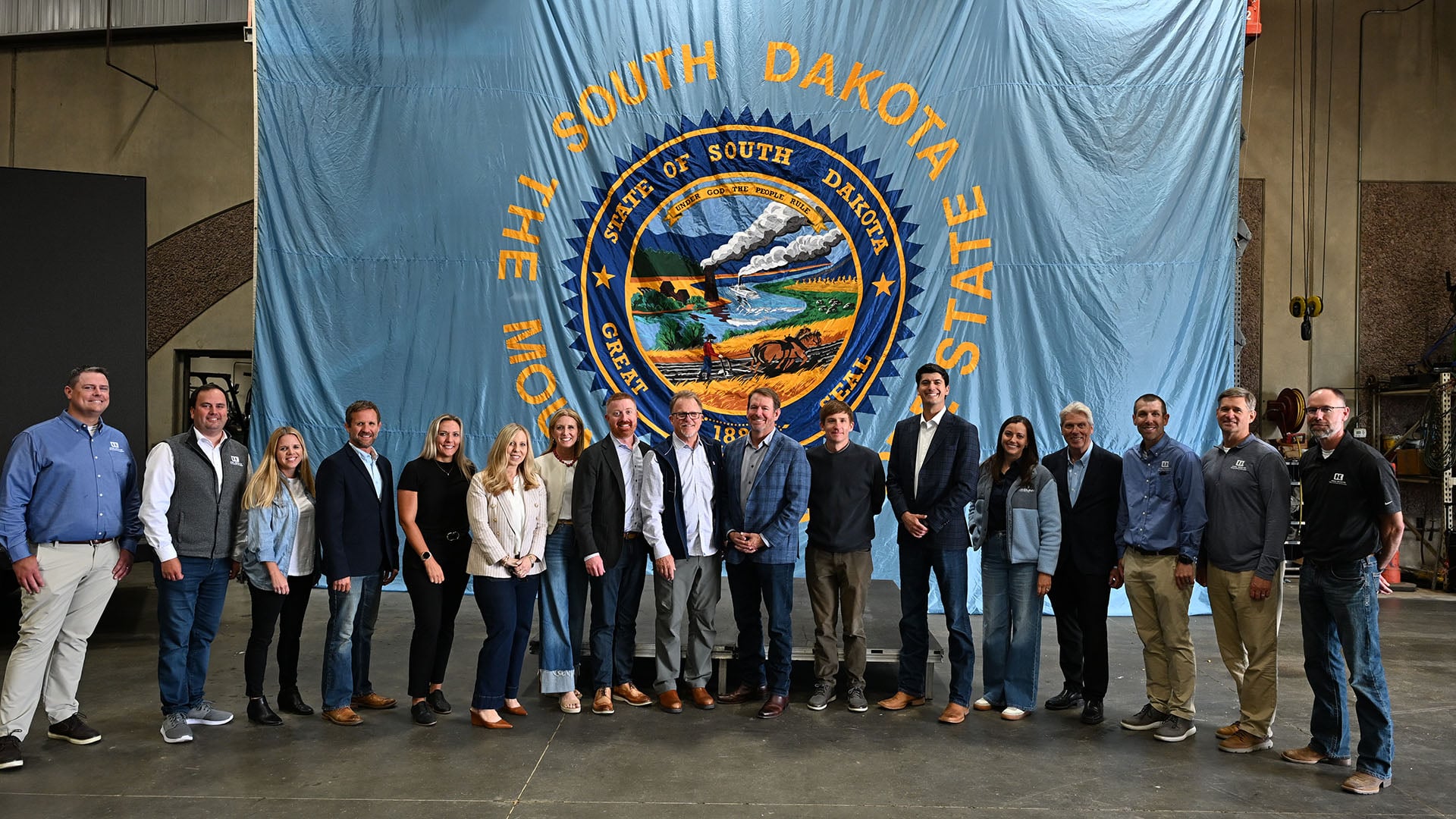 Group of professionals posing side by side indoors in front of a large blue banner with the South Dakota state seal image in the center and bold yellow lettering above and around it.