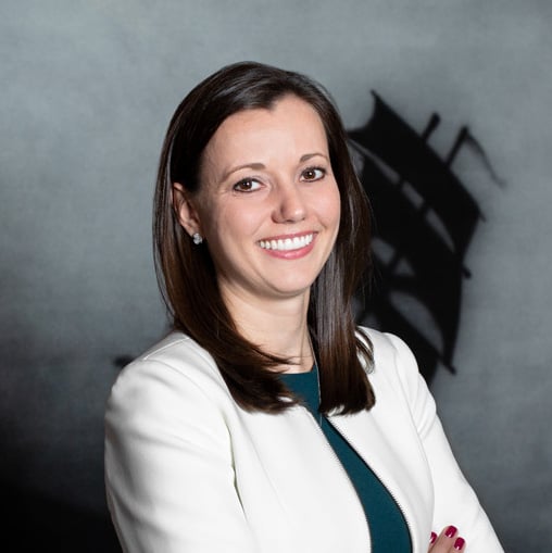 Professional headshot of a smiling woman with dark hair, white blazer, and teal blouse, posed against a neutral backdrop