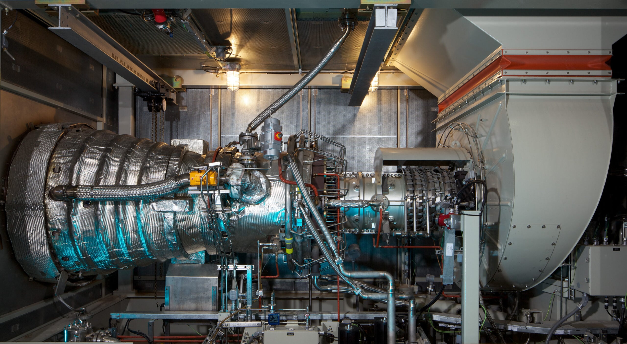Jacobs Engineering OU Power Plant 4302012 | JE Dunn Construction Large industrial turbine or jet engine setup with insulated ducts, pipes, and metallic framework in a dimly lit mechanical room; complex machinery visible for testing or maintenance purposes.