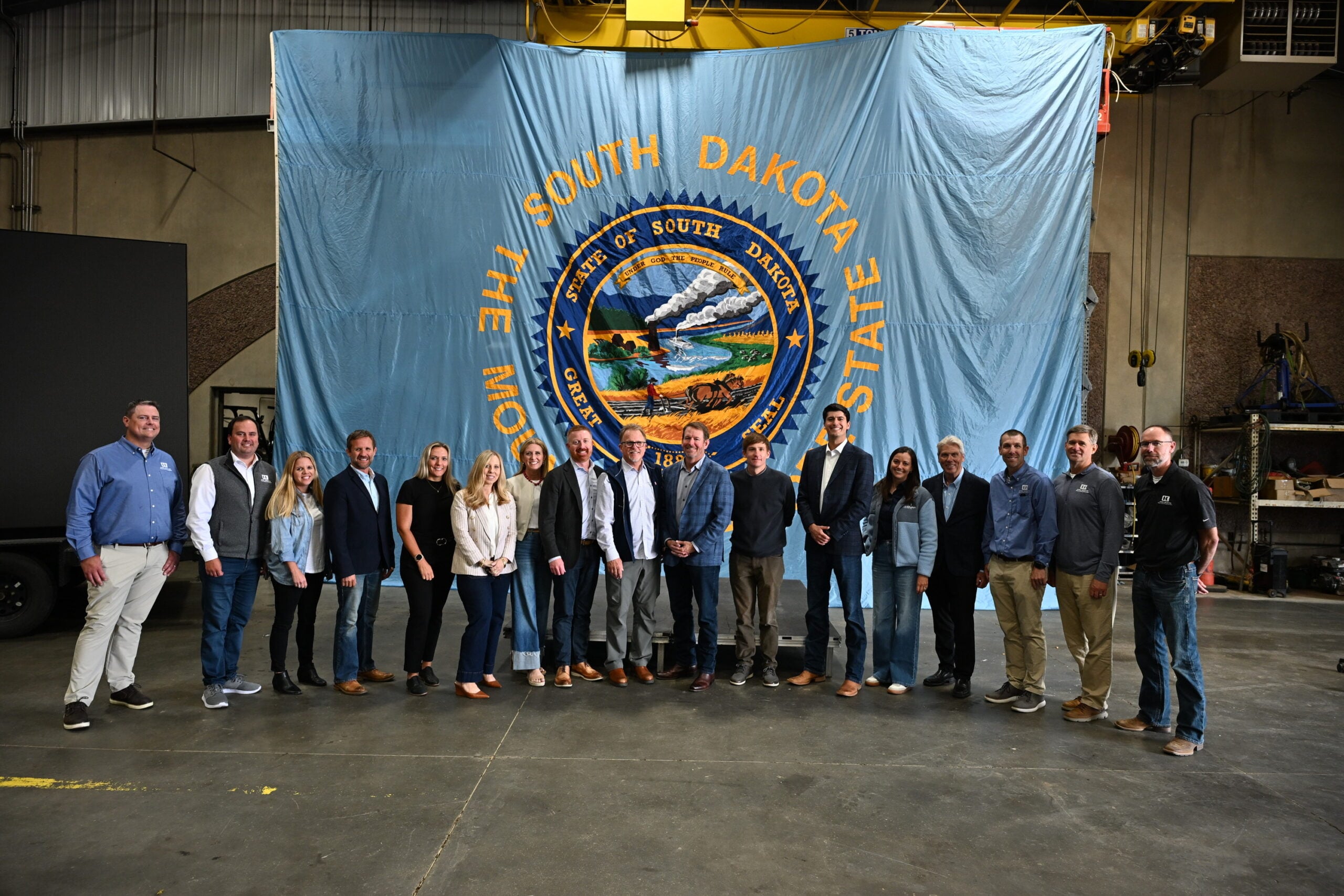 Group of about 20 people posing in a workshop in front of a large blue banner showing the South Dakota state seal.