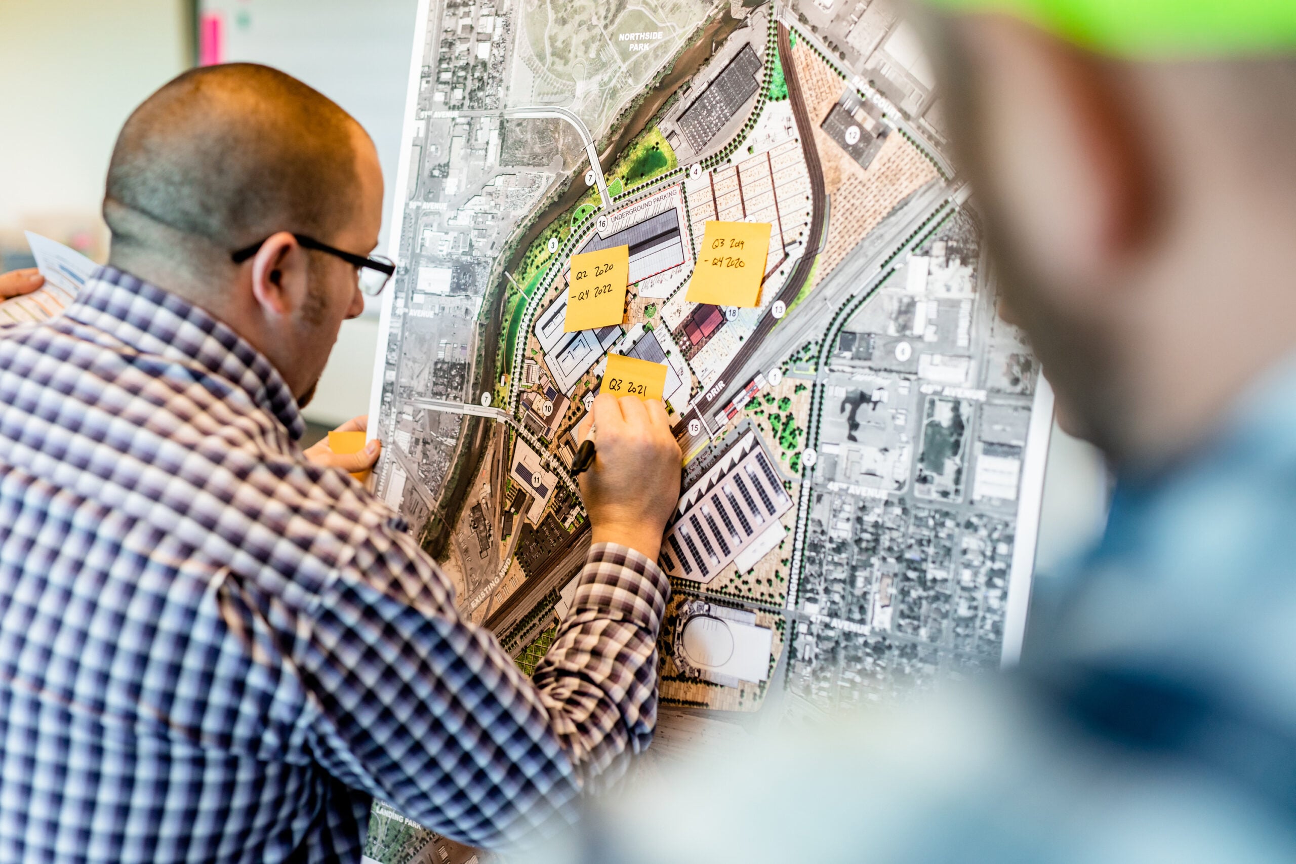 | JE Dunn Construction Two people review a large urban planning map with yellow sticky notes marking project phases during a planning meeting