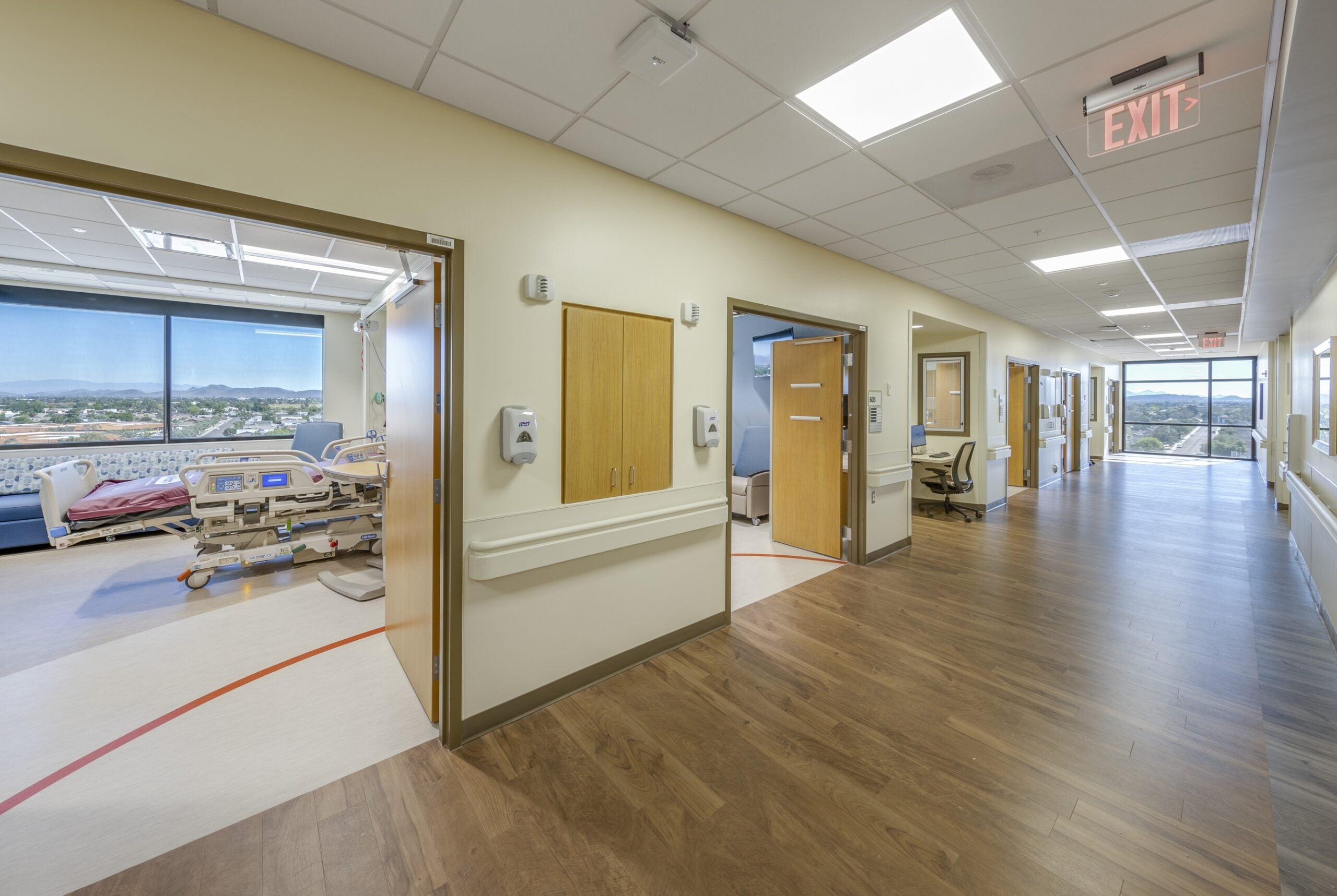 Banner Health Thunderbird Medical Center 4-6th Floor Buildout_I_H_Patient Cooridor Rooms | JE Dunn Construction Hospital hallway in a medical facility with patient rooms on the left and an exit sign ahead in a bright, wide corridor