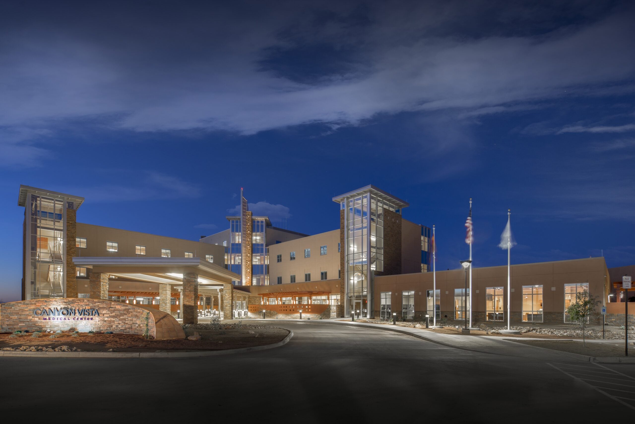 Canyon Vista Medical Center_E_H_Entrance | JE Dunn Construction Nighttime exterior of Canyon Vista Medical Center with a lit entrance, brick pillars, and glass towers against a deep blue sky.