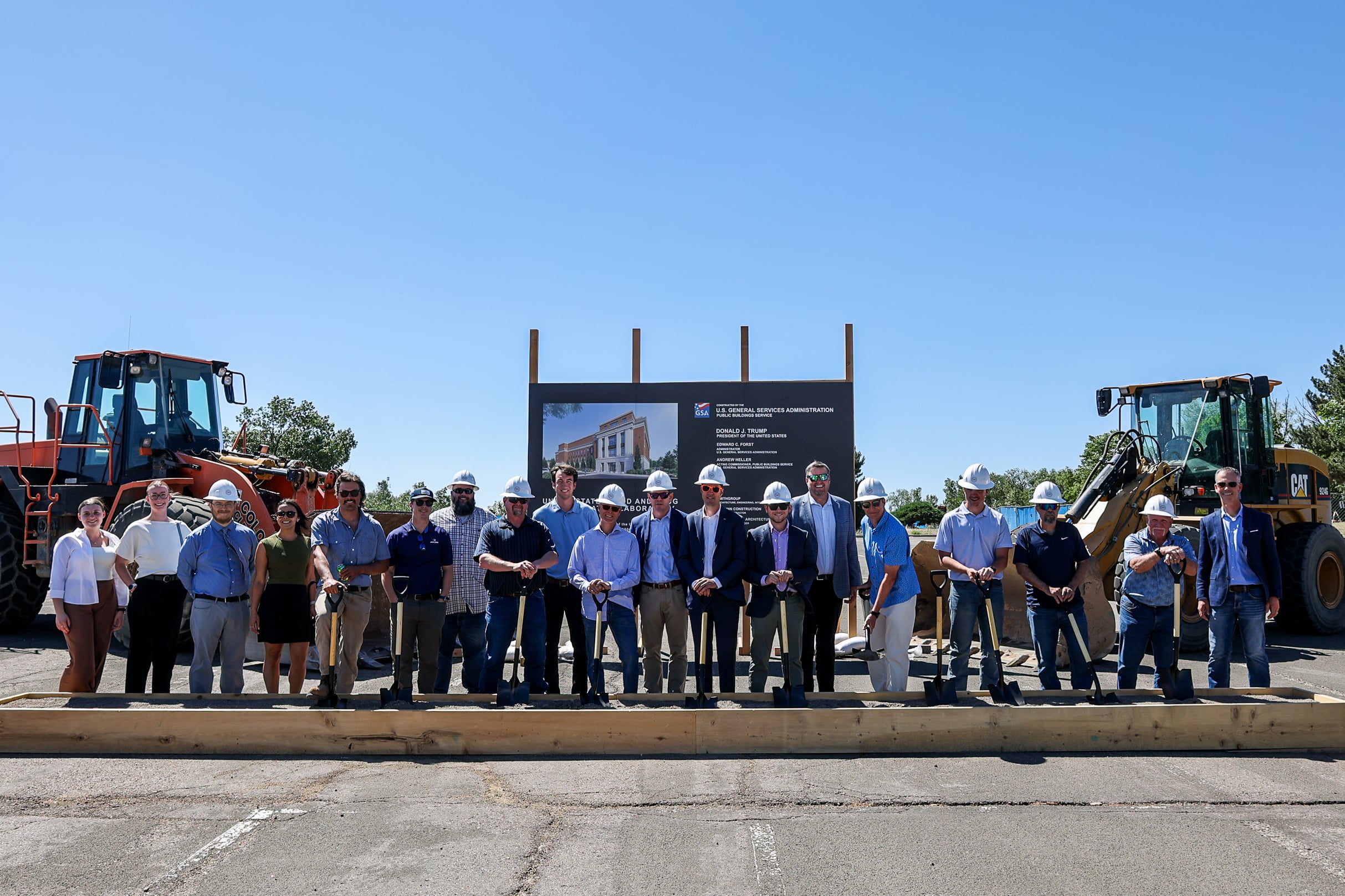 Group of professionals wearing hard hats posing at a construction site during a groundbreaking ceremony, with equipment in the background.