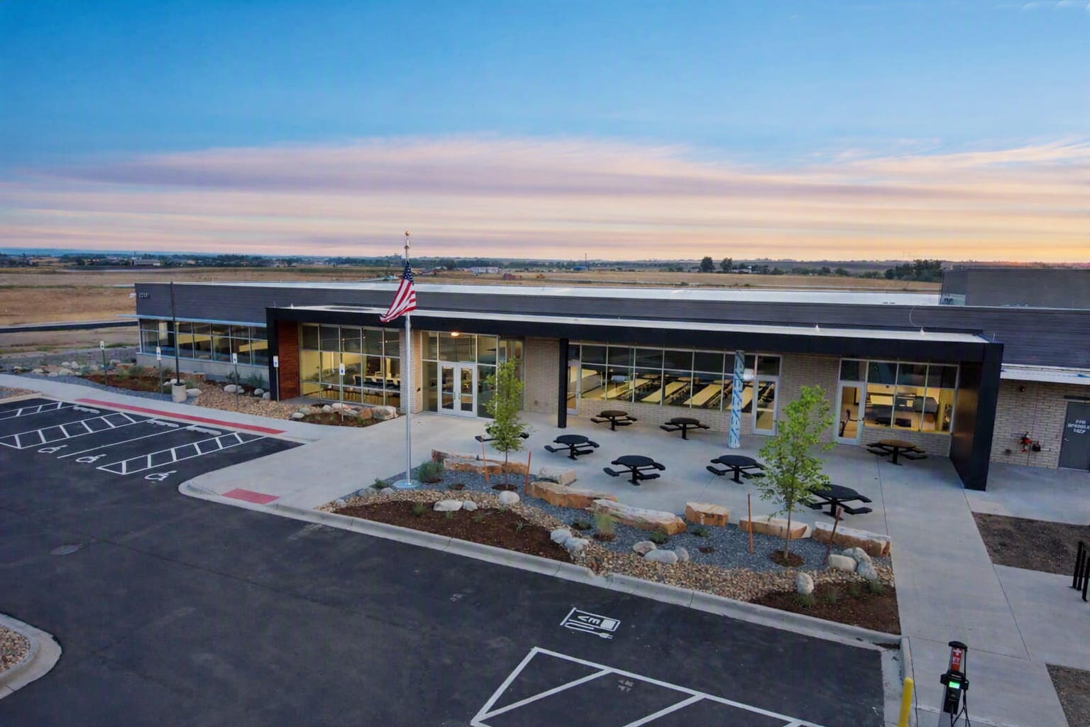 LCESAir015 | JE Dunn Construction A modern single-story building with large glass windows, American flag on a flagpole, and circular outdoor seating near landscaped beds.