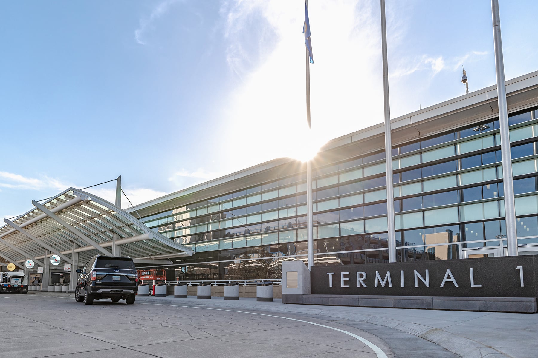 Airport terminal exterior with glass façade, flagpoles, and a black SUV near the entrance; sign reads TERMINAL 1 with blue sky above.