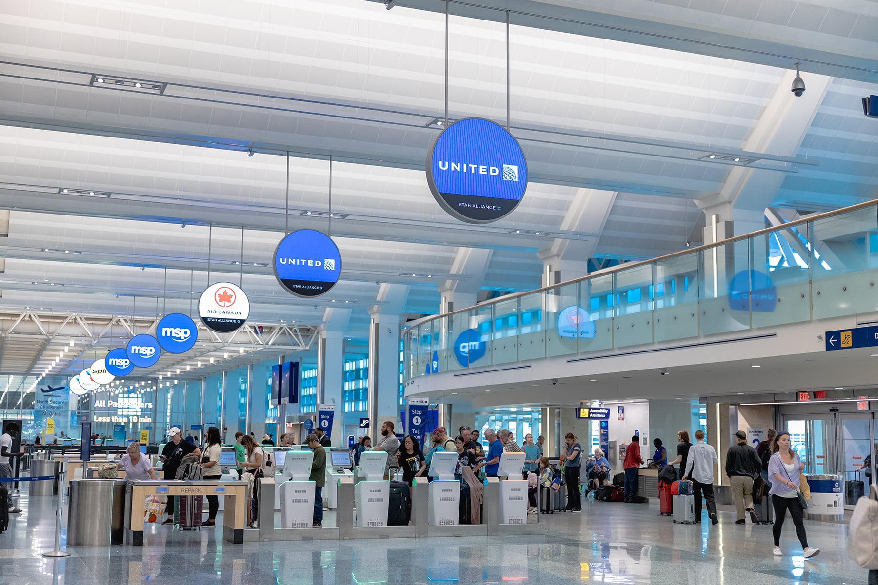 Airport check-in area with counters and blue circular signs (United, MSP, Air Canada) above; travelers with luggage in line and staff at desks.