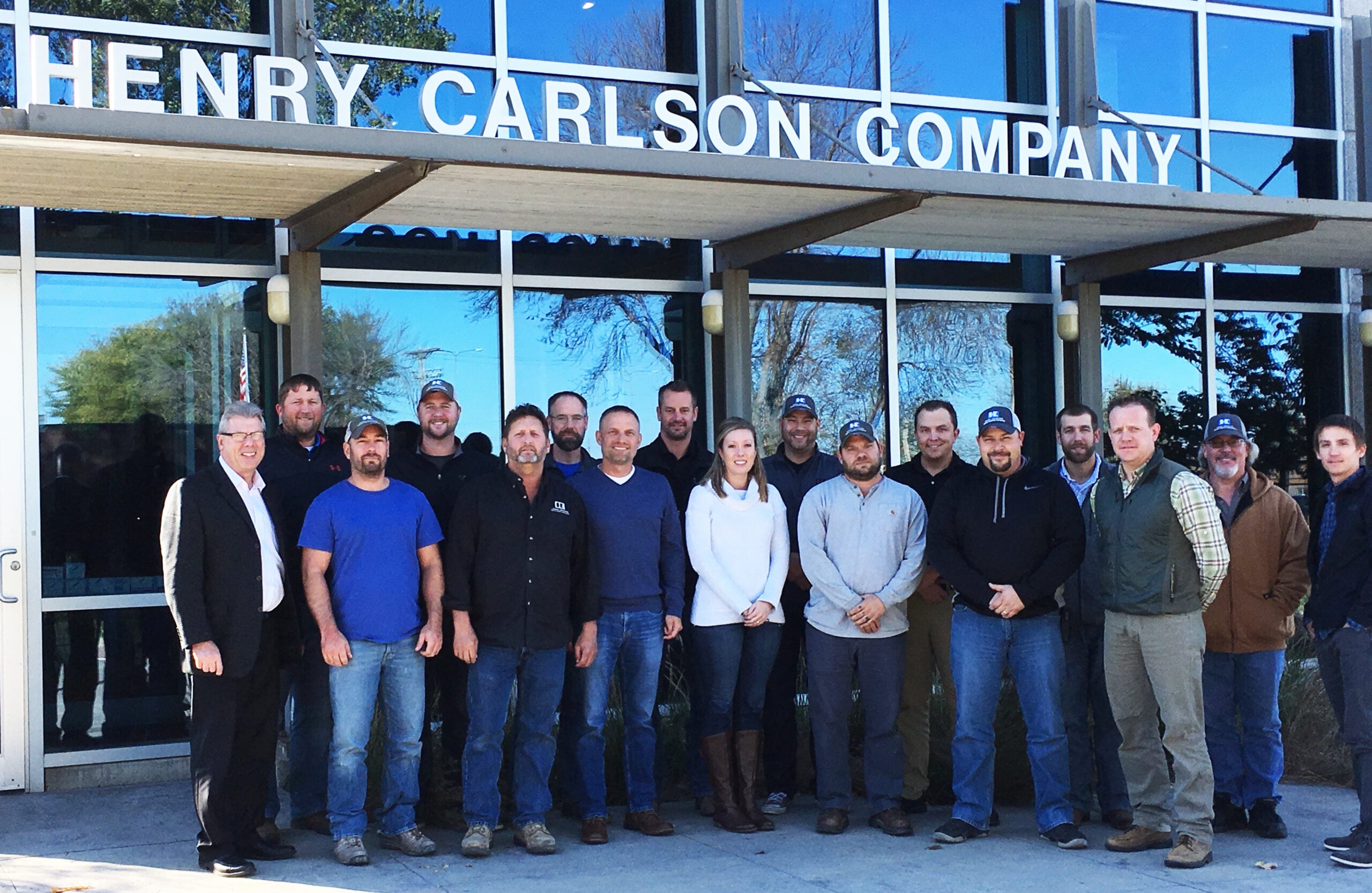 Group of about 20 employees posing for a company photo in front of a glass-walled Henry Carlson Company building entrance.