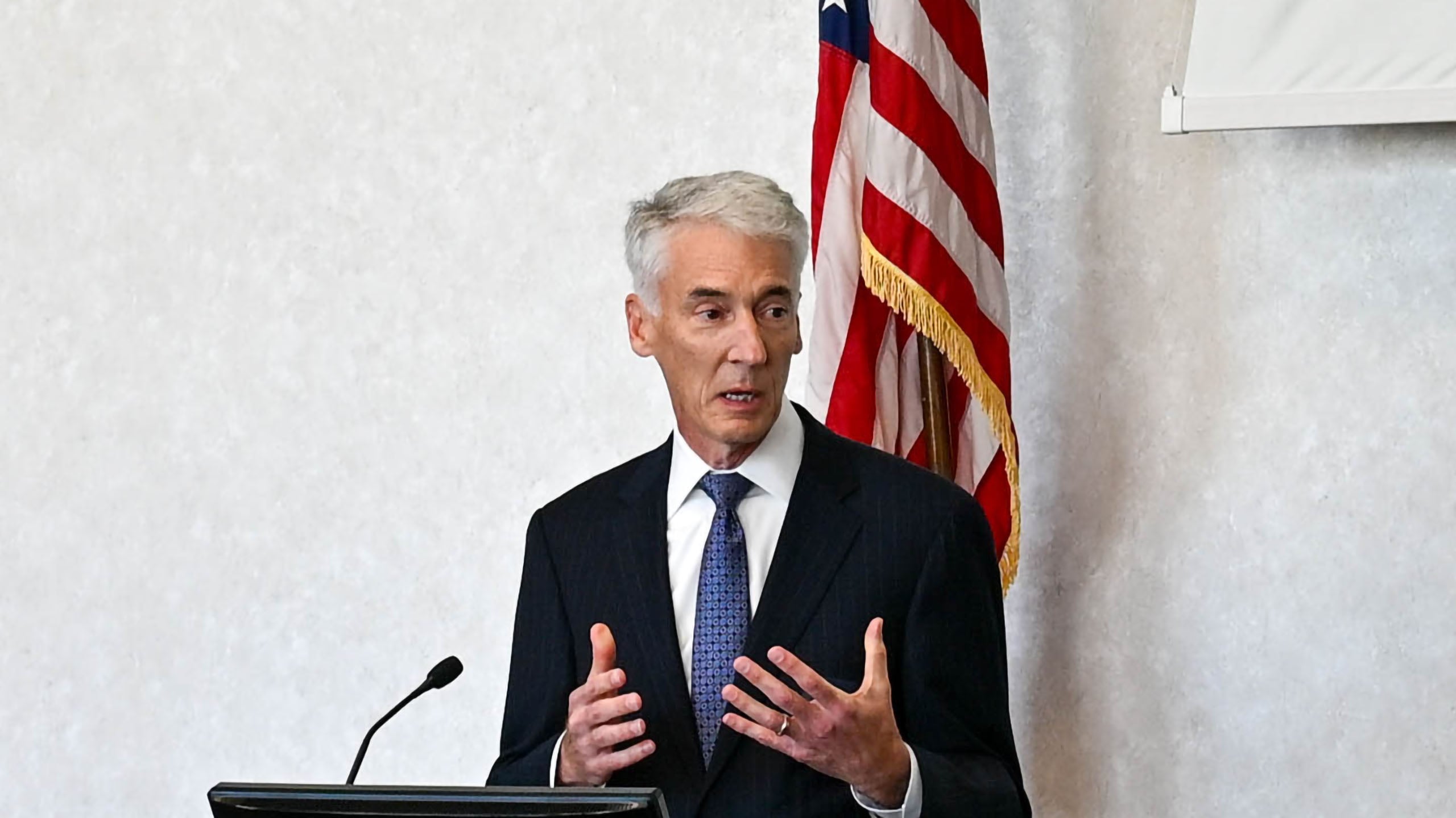 Man in a dark suit and tie speaks at a podium, with an American flag in the background.
